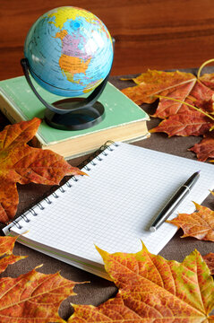 Autumn background with education, back to school, university studying concept. White checkered notebook, vintage ink pen, worn book and a globe on the table among autumn leaves, free space for text