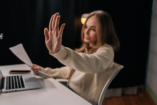 Portrait of serious confident businesswoman focused at desk holding papers and extends hand in stop gesture to signal refusal of interruption working on laptop in dark modern office room.