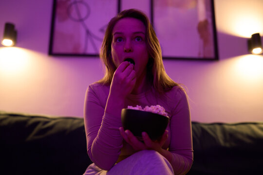 Female sitting on sofa eating popcorn while watching screen indoors