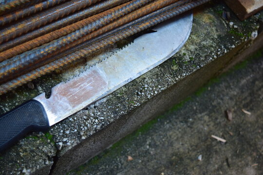 A sharp, shiny machete of a peasant, with a peculiar shape, sits atop a wall next to some rods.