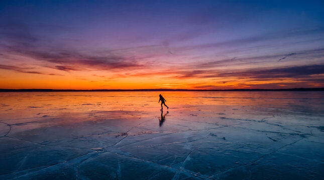 Silhouetted person walking on frozen lake surface during sunset with vibrant sky and reflections