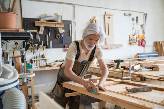 Professional craftsman working on restoration of antique wooden door at workbench in carpentry, determined man woodworker smoothing and sanding wood surface with sandpaper at worktable in workshop.