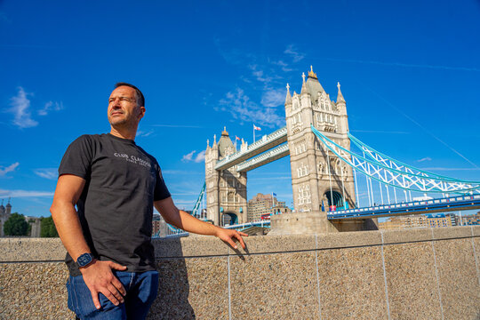 A white European man in a black T-shirt posing on a wall with London Bridge in the background.