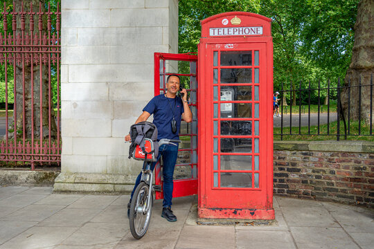 A white European man riding a bicycle, wearing a telephone headset, outside a phone booth in London.