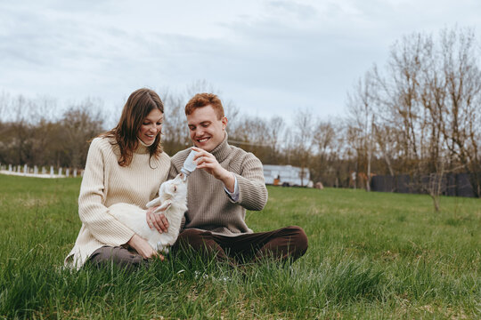 Cheerful couple outdoors, interacting with a friendly goat kid, sitting on grass in a sunny meadow, warm sweaters, relaxed mood, natural colors, peaceful bonding moment amid open countryside