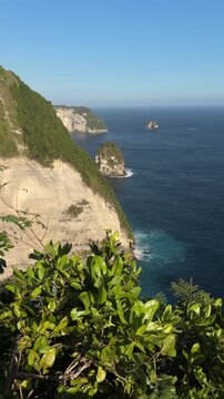 A scenic coastal view featuring steep cliffs, lush greenery, and a clear blue ocean. The horizon meets the sky under bright sunlight.