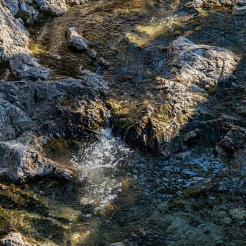 Narrow stream water moving between rocks, creating bright reflections, dark pools, and a calm natural rhythm.