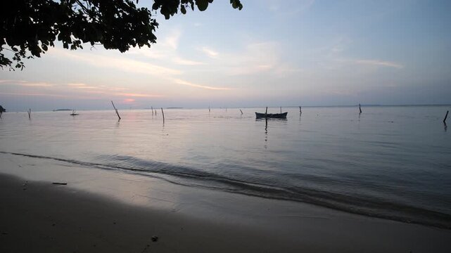 Peaceful tropical beach at sunset in Karimunjawa National Park Indonesia with small fishing boat anchored on calm sea, framed by coastal trees beneath pastel evening sky