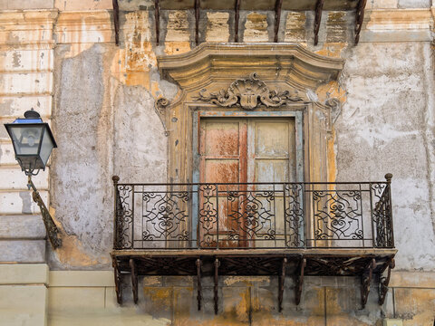 A classic architectural shot of a traditional Sicilian wrought-iron balcony in Palermo, featuring a weathered Baroque window frame and an antique street lantern against a textured stucco wall.