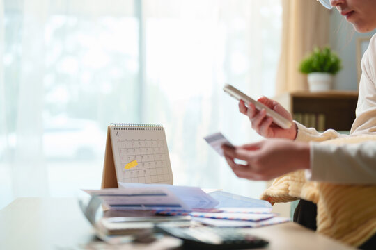 Young woman using smartphone and credit card to pay bills and manage monthly budget with calendar and documents at home. Personal finance, online banking, budgeting and financial planning