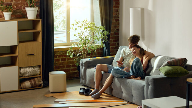 Young couple browsing renovation products on smartphone at home. Concept of mobile shopping and shared decisions in e-commerce, flooring retail and relocation campaigns.