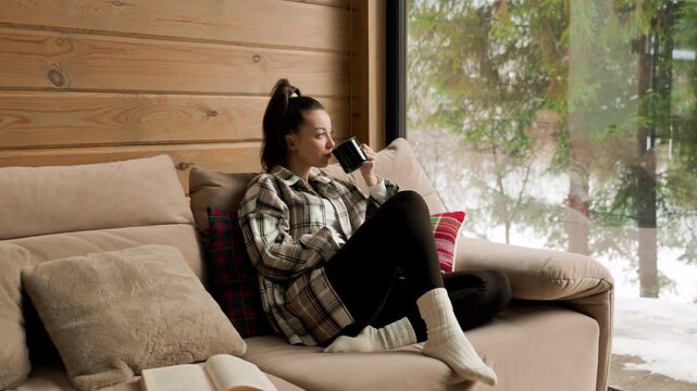 Young woman drinking hot coffee while relaxing on cozy sofa in modern wooden cabin with panoramic window overlooking snowy winter forest peaceful hygge lifestyle concept