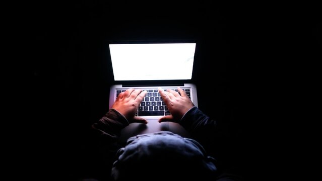 Hacker typing on laptop keyboard in dark room, high contrast digital security photo