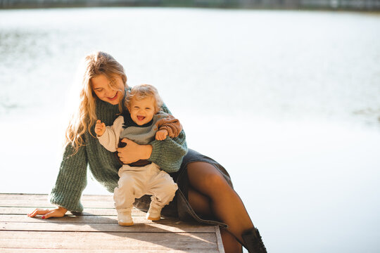 Smiling mother holding baby 1-2 year old wearing knitted sweater sitting on wooden pier over lake water outdoors. Motherhood.
