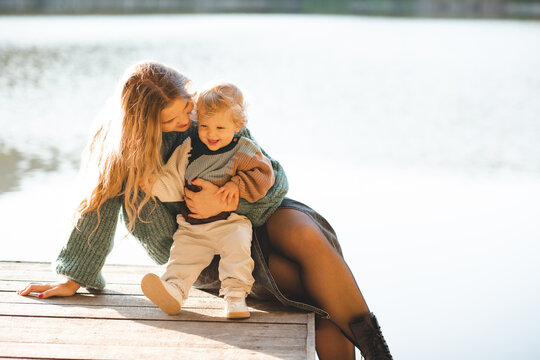Smiling mother holding baby 1-2 year old wearing knitted sweater sitting on wooden pier over lake water outdoors. Motherhood.