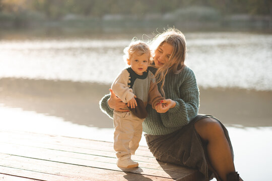 Cute mother holding baby 1-2 year old wearing knitted sweater sitting on wooden pier over lake water outdoors. Motherhood.