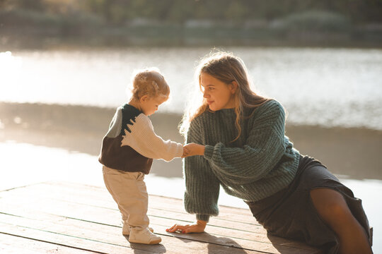 Stylish mother holding baby 1-2 year old wearing knitted sweater sitting on wooden pier over lake water outdoors. Motherhood.