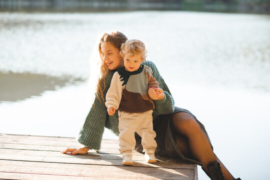 Cute mother holding baby 1-2 year old wearing knitted sweater sitting on wooden pier over lake water outdoors. Motherhood.