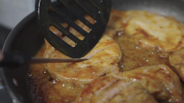 Close-up of marinated pork steaks cooking in savory sauce in a frying pan on a gas stove. A person uses a black spatula to flip the meat. Home cooking and delicious dinner food preparation.