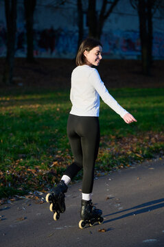 Back view of young smiling woman in rollerblades moving along asphalt track, focused on ride in peaceful park surroundings. Good for mobility, recreation, outdoor sport or wellness campaigns