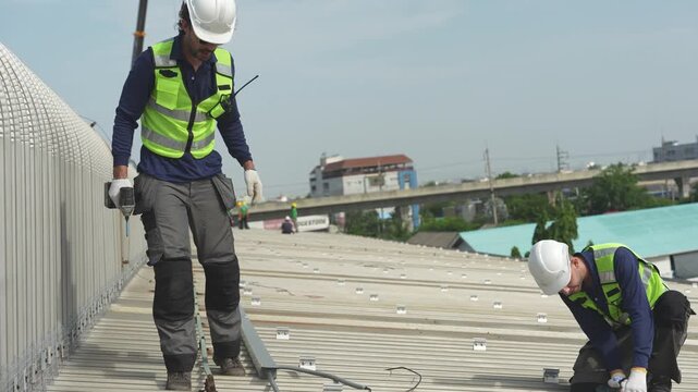 Two engineers kneel on the bare metal roof fastening mounting rails, preparing the ground for the first solar modules of the industrial installation, filmed as part of a commercial clean