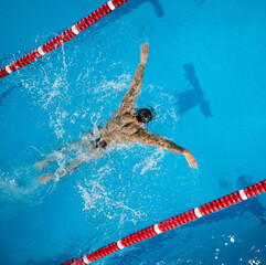 Fototapeta premium Top view of tattooed male swimmer performing butterfly stroke in pool lane with dynamic splash. Concept of fitness lifestyle, perseverance, discipline, health, active living and goal achievement