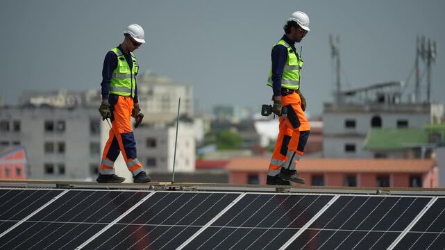 Two inspectors in yellow hi vis jackets walk the ridge line above the array, low rise city rooftops spreading into the distance behind them, filmed while a new industrial rooftop