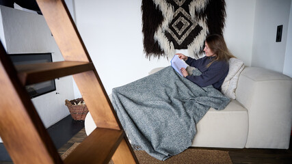 Obraz premium Woman reading book on sofa wrapped in blanket, relaxing at home near fireplace, cozy interior with warm atmosphere, leisure time and comfortable lifestyle concept