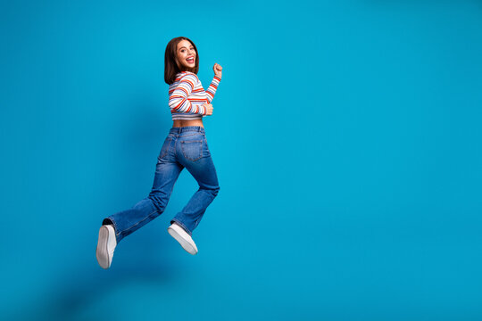 Young chic woman jumps in bright blue studio wearing striped shirt and jeans smiling for lifestyle and fashion advertisement
