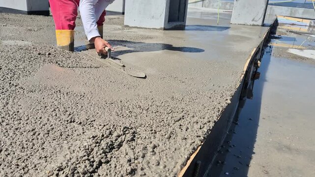 Construction worker smoothing fresh concrete with hand trowel standing in wet concrete on a sunny day.
