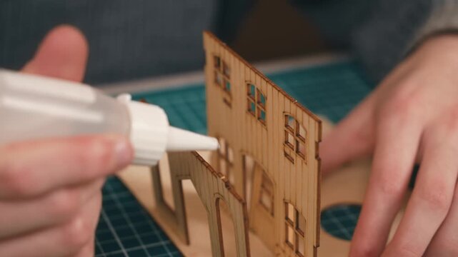Applying adhesive to wooden house model elements during intricate assembly. Maker workshop process featuring laser cut plywood parts and handcrafted structural detailing. Building tomorrow home vision