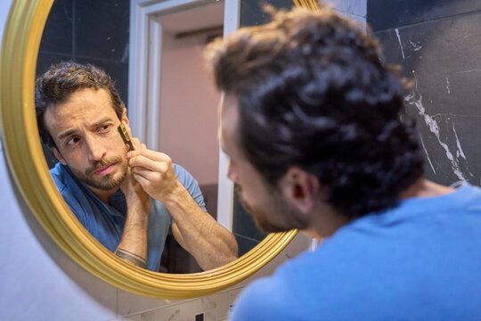 Young man grooming beard with trimmer in bathroom mirror