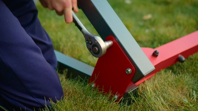 Outdoor worker tightening bolt with ratchet wrench on red bracket and gray metal frame. DIY assembly repair task on grass with socket tool and hardware connection. Construction installation concept