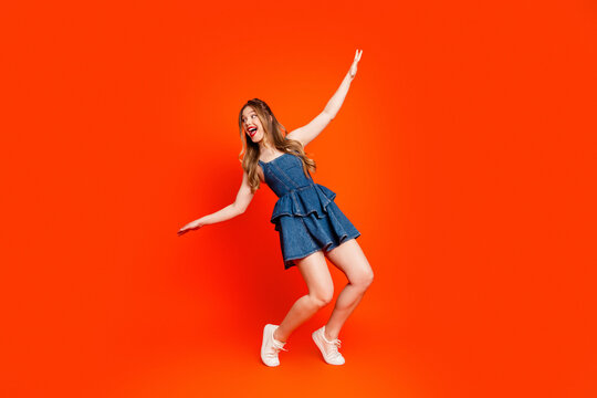 Young woman in denim dress dancing and laughing on bright orange background expressing playful joy and confident summer style