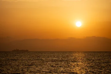 A golden sunset casts warm light over calm ocean waters while a cargo ship sails as a dark silhouette on the distant horizon, creating a peaceful, dramatic coastal seascape. © Andrea Izzotti