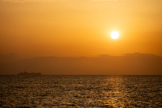 A golden sunset casts warm light over calm ocean waters while a cargo ship sails as a dark silhouette on the distant horizon, creating a peaceful, dramatic coastal seascape.