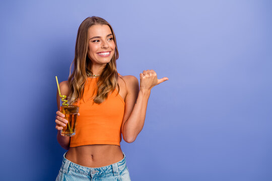 Young chic woman in an orange singlet holding a drink and smiling against a purple background for lifestyle fashion and promotional use