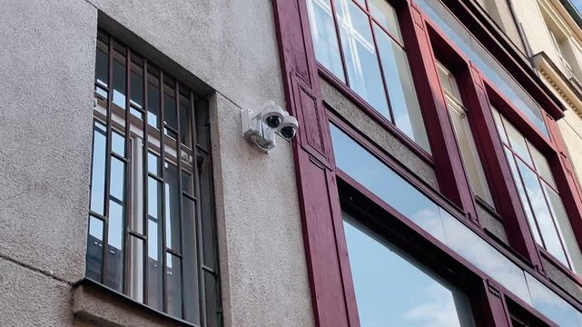 neutral, static urban street scene with building facade and security cameras in a low angle close-up view, showcasing concrete, glass, and metal materials under beige and red tones.