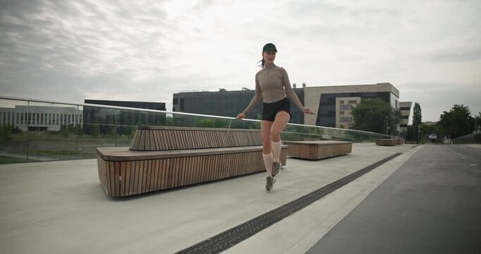 A young beautiful girl in sportswear and a cap does exercises with a skipping rope and plays sports in an urban setting on a bridge with a pedestrian area and benches against the backdrop of new concr