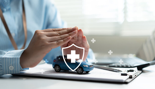 Businesswoman hands protecting car with shield and cross icon at desk. Concept of auto insurance, vehicle safety, medical coverage and financial protection care.