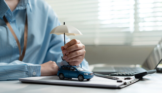 Businesswoman holding small white umbrella over car at office desk. Concept of auto insurance policy, vehicle protection, safety coverage and financial security.