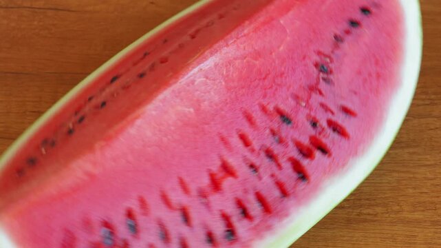 A slice of ripe red watermelon rotating on a textured wooden table surface with a static camera view. High speed fps footage for summer food commercial, organic fruit and refreshing dessert video. 