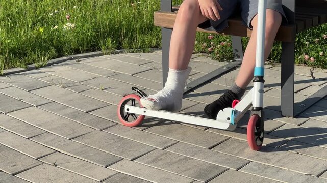 A kid with a broken leg in a cast sits on a bench with a scooter nearby on a sunny day, childhood accident, recovery process, summer leisure.