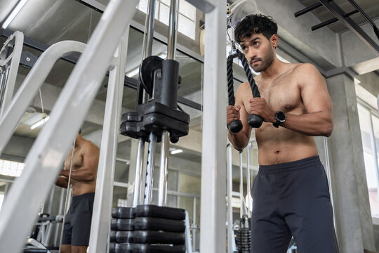 Focused muscular Asian man performing triceps pushdown exercises on a cable machine during an intense strength training workout inside a modern fitness center building healthy endurance.