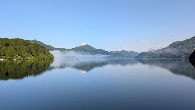 the calm, smooth waters of Lake Ashi with morning fog on the surface and fishing boats surrounded by forested mountains and overlooking Mount Fuji with its snow-capped peak against a blue sky