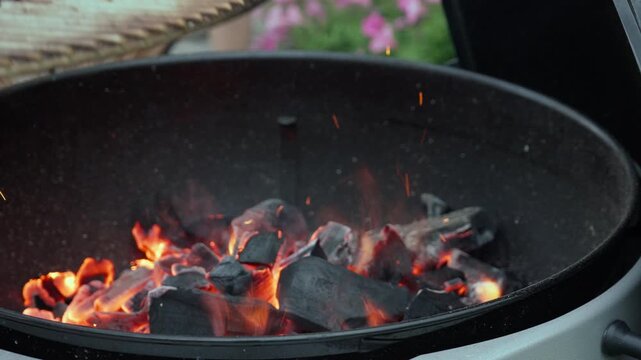 A person wearing a heat resistant glove places a steel grate over burning charcoal. Hot glowing coals flare with sparks on an outdoor grill. Preparing equipment for summer barbecue cookout.