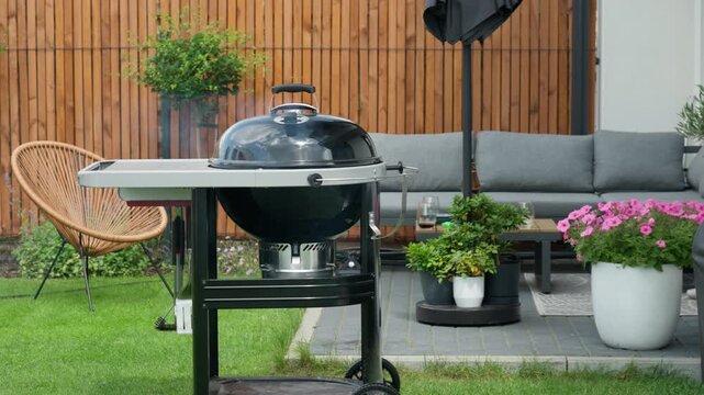 A closed kettle charcoal grill steams on a modern backyard patio lawn. Food cooks slowly inside during a summer barbecue. Perfect setting for outdoor family dining and weekend relaxation.