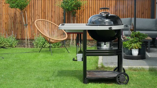A closed charcoal grill steams on a green lawn in a modern backyard patio terrace. Light smoke drifts from the kettle grill during a summer barbecue cookout. Great setting for outdoor dining.