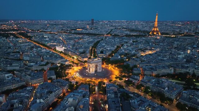 Aerial night view of the famous landmark Triumphal Arch or Arc de Triomphe and Eiffel Tower surrounded by busy streets and lush greenery in the heart of the city Paris, France. Drone orbiting flight