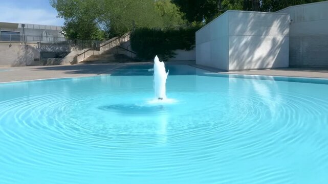 serene fountain spraying water in an outdoor pool area with large rocks and trees in the background. relaxing scene with blue and green tones and concrete and stone materials.